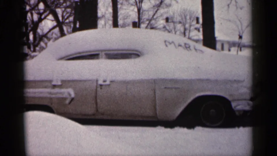 Closer 1961 Minnesota 8mm view of a vintage car covered in snow, framed by a driveway snowbank and trees.