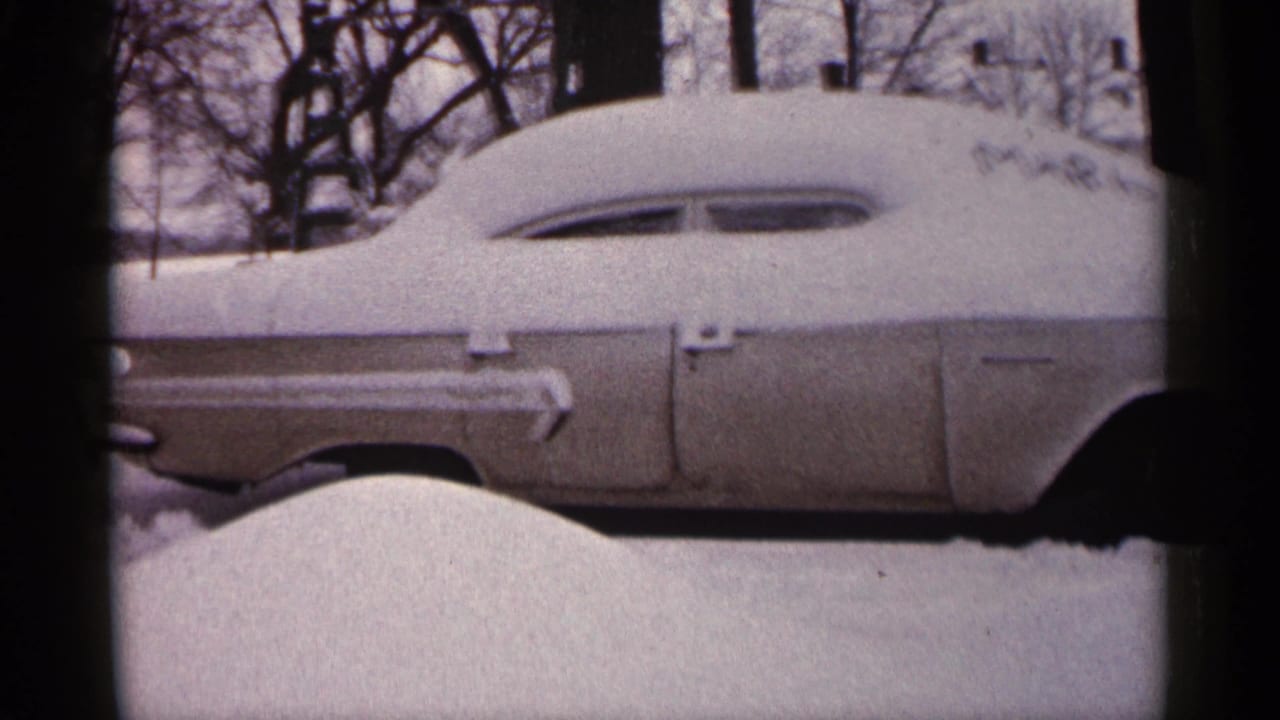 Snow-covered older car parked outdoors beside bare winter trees.