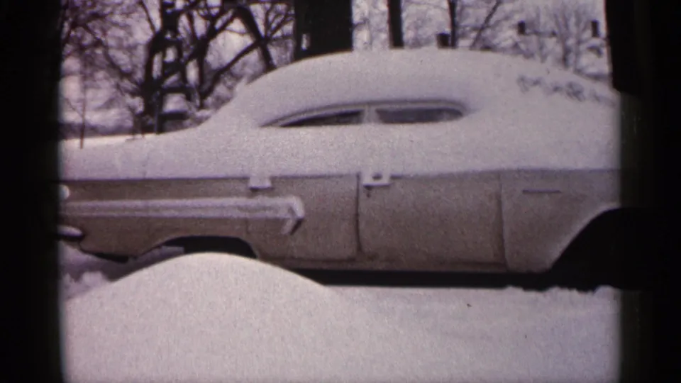 1961 Minnesota home-movie frame showing an older car partly buried under snow, with bare trees and deep snow in the background.