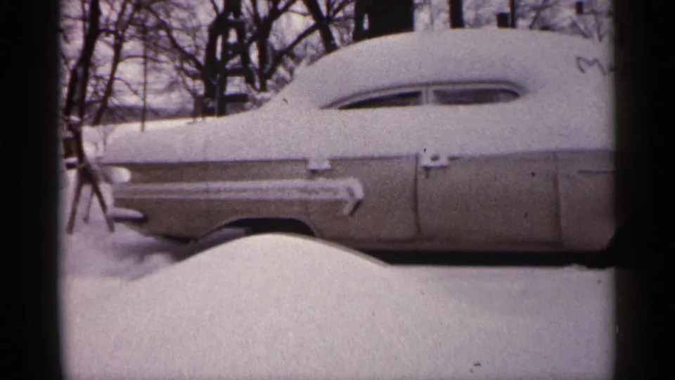 Side view of a snow-covered older car in a 1961 Minnesota winter scene, with deep snow and leafless trees behind it.