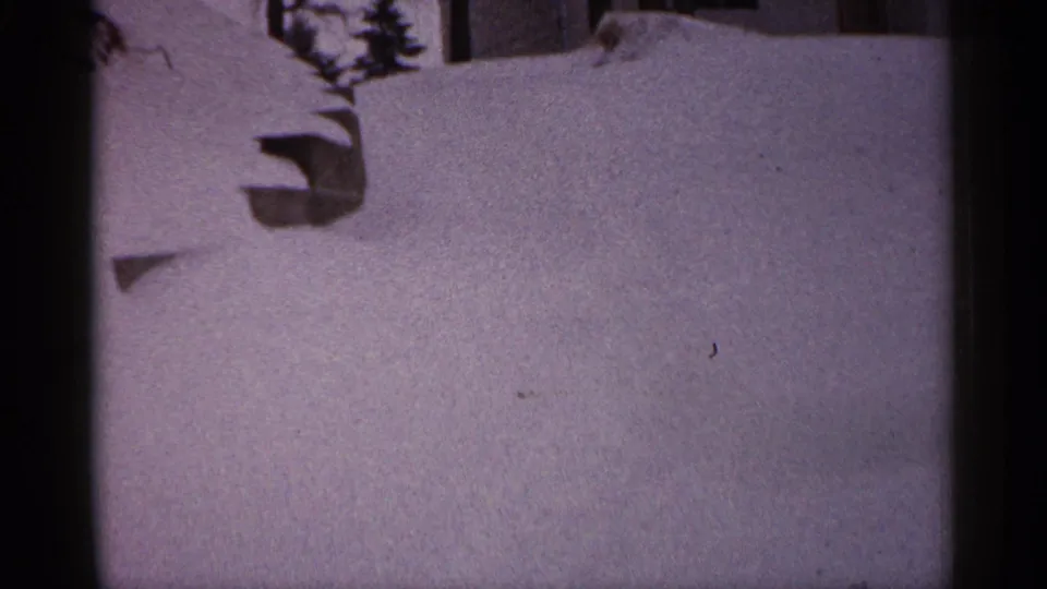 1961 Minnesota winter frame with deep snow across a residential yard, a low object protruding from the snow, and partial building detail above.