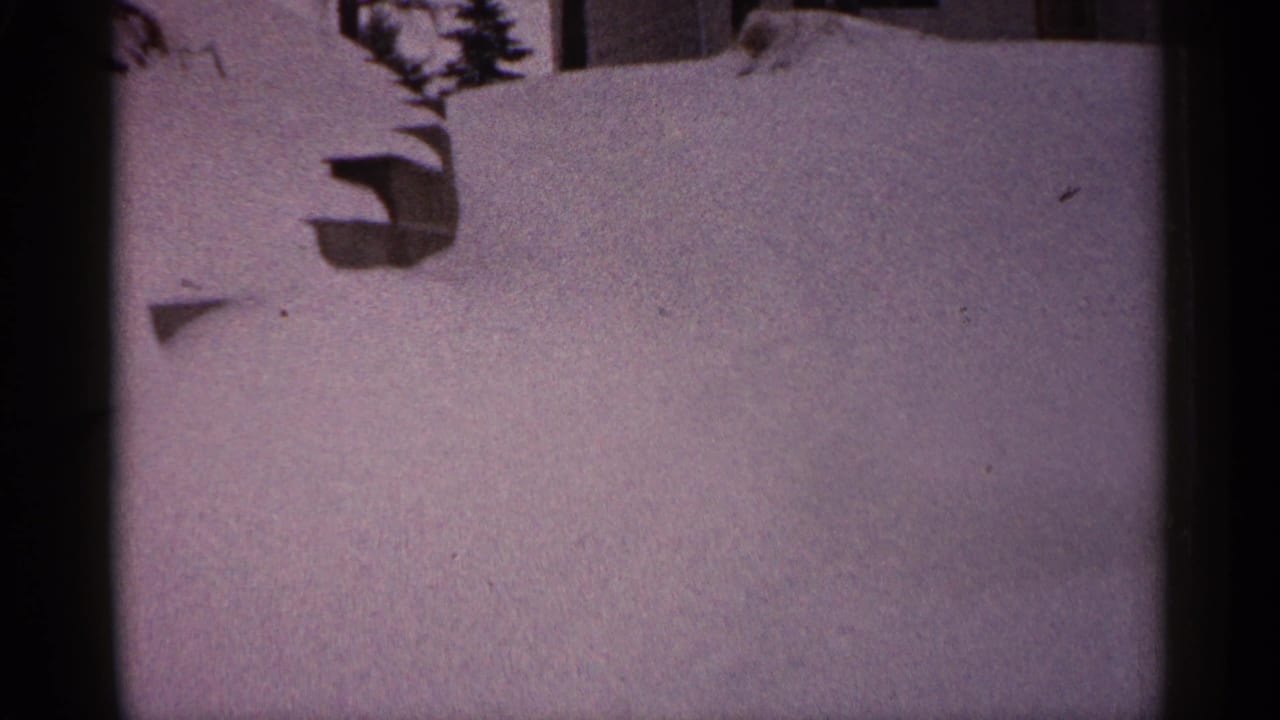 1961 Minnesota home-movie frame of a snow-covered residential area with a wall, branch shadows, and uneven snow surface.