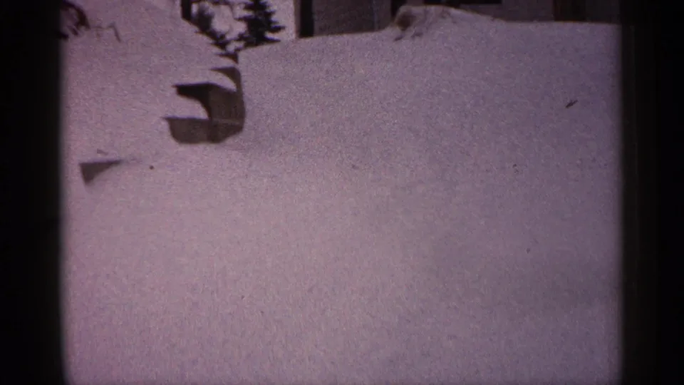 1961 Minnesota home-movie frame of a snow-covered residential area with a wall, branch shadows, and uneven snow surface.