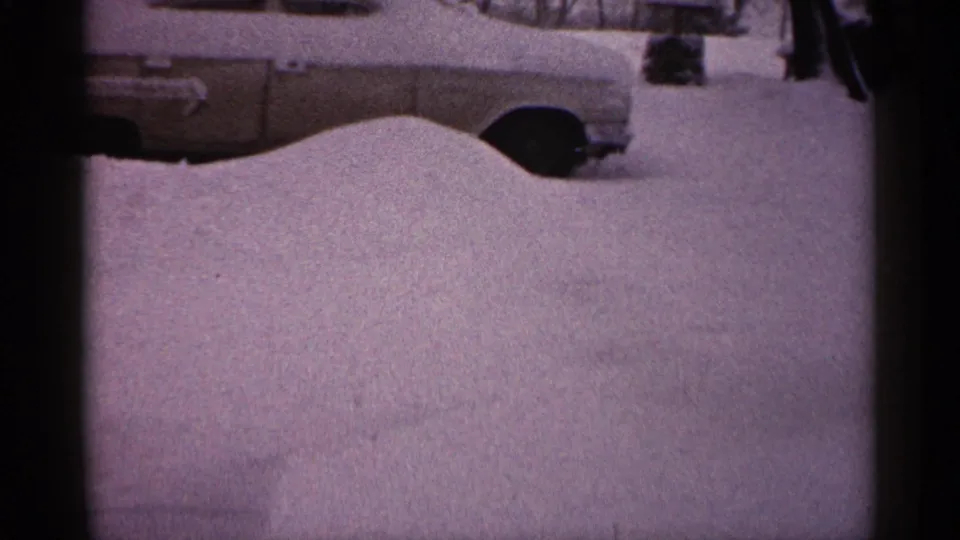 A grainy 1961 Minnesota winter frame shows the side of a classic car emerging above a deep snow drift.