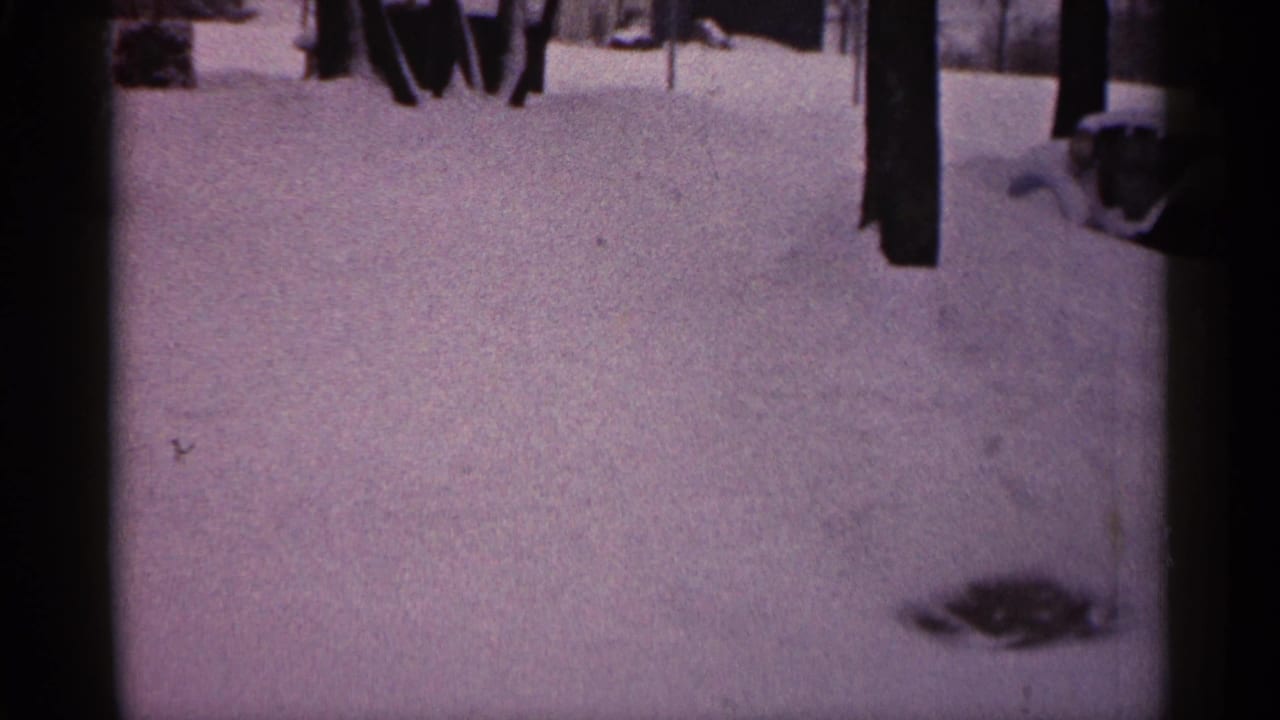 1961 Minnesota home-movie frame showing deep snow, tree trunks, partial adult figures, and a partly buried car near a house.