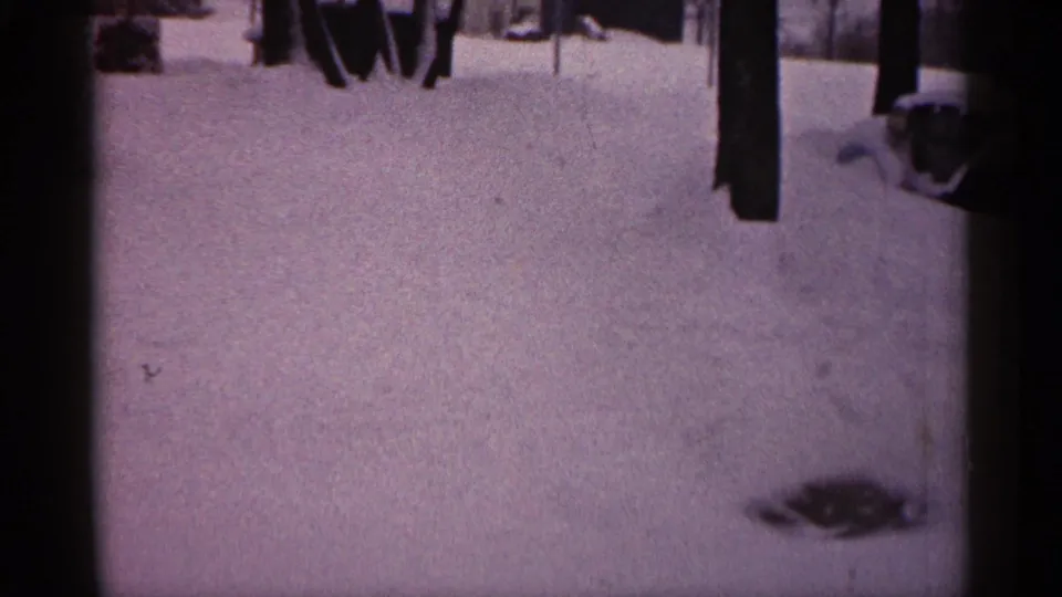 1961 Minnesota home-movie frame showing deep snow, tree trunks, partial adult figures, and a partly buried car near a house.