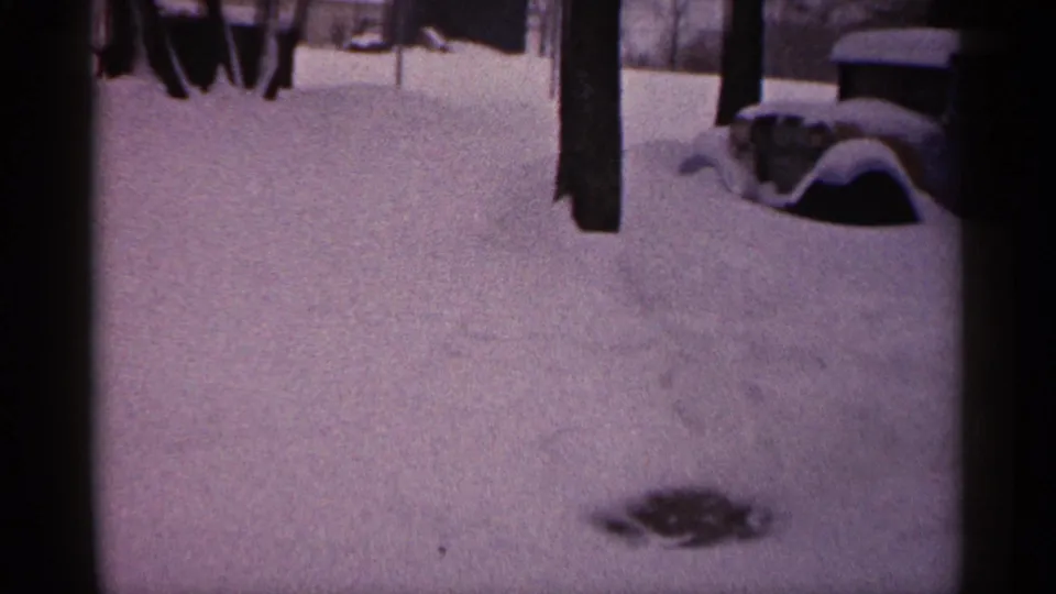 1961 Minnesota winter frame with accumulated snow across a yard, partial adults near the top edge, and a snow-covered car at right.