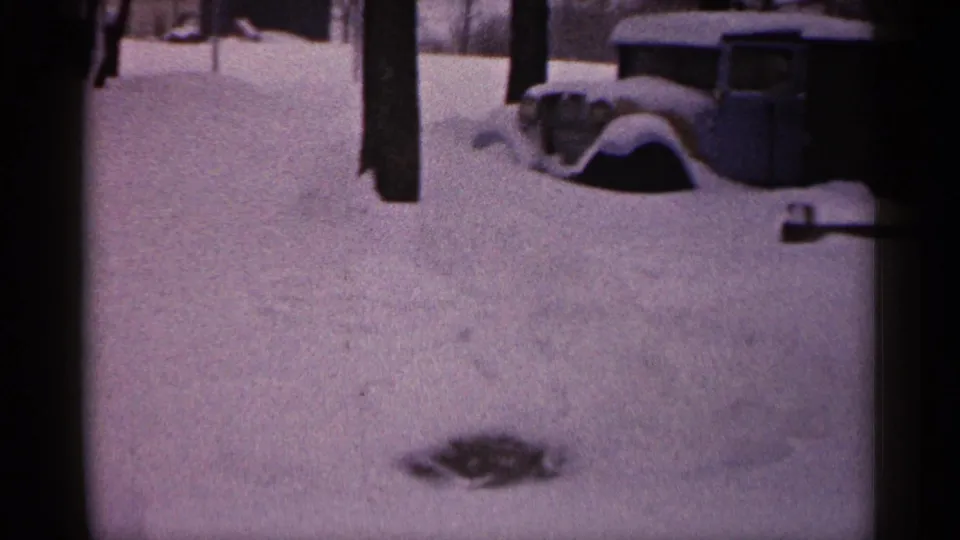 1961 Minnesota 8mm frame showing a quiet snowy yard, tree trunks, a buried car, and the edge of a home or porch.