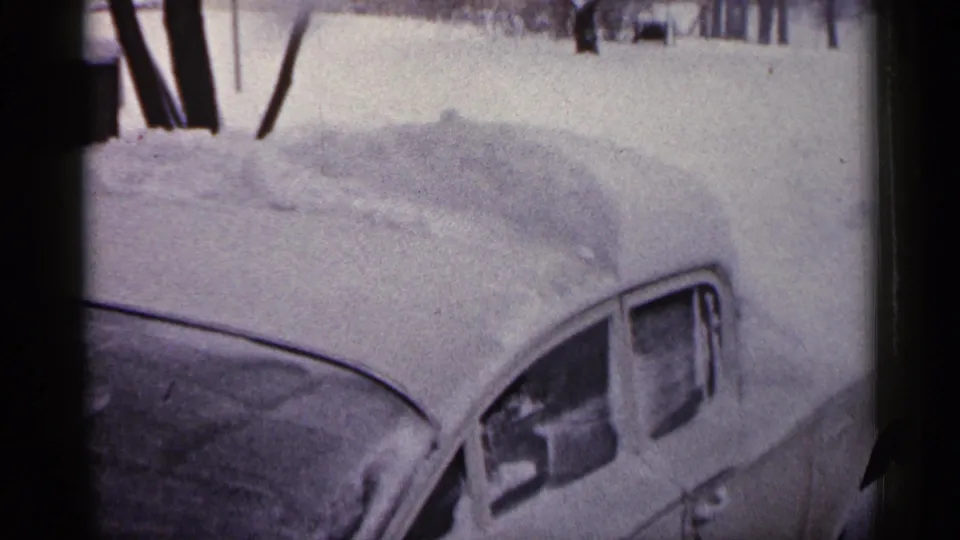 1961 Minnesota 8mm frame with a close view of an older parked car buried under snow, with trees in the background.