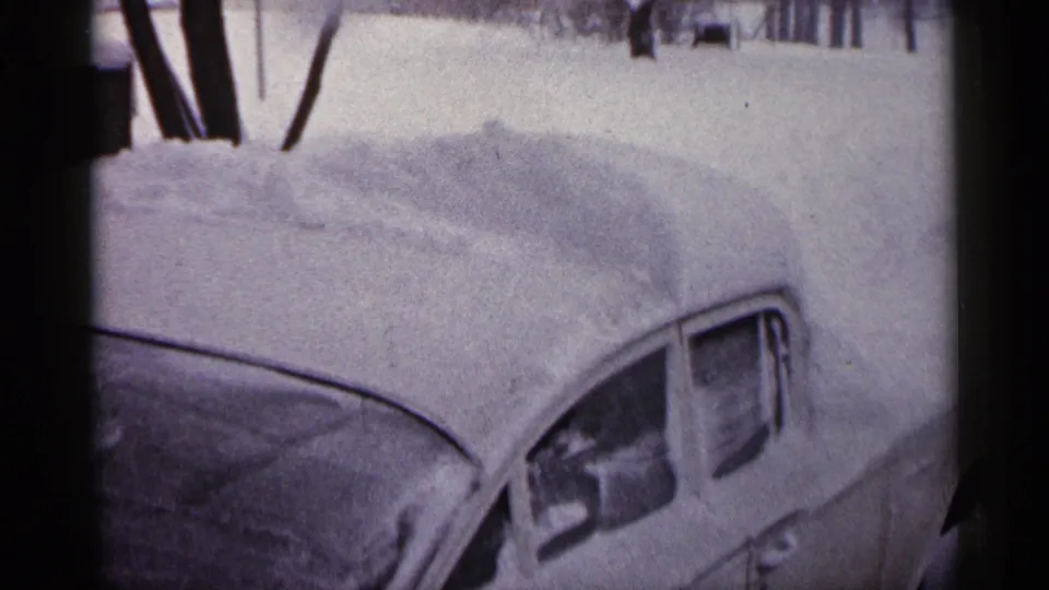 1961 Minnesota home-movie frame showing a classic parked car buried under snow near bare trees and deep roadside accumulation.