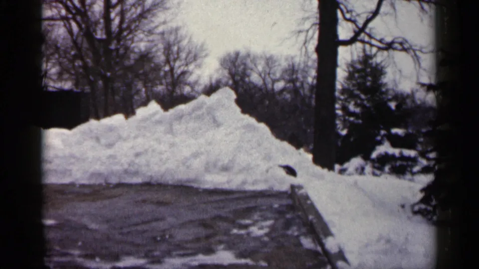 1961 Minnesota winter frame showing a large plowed snow pile along pavement, with leafless trees and a dark structure in the background.