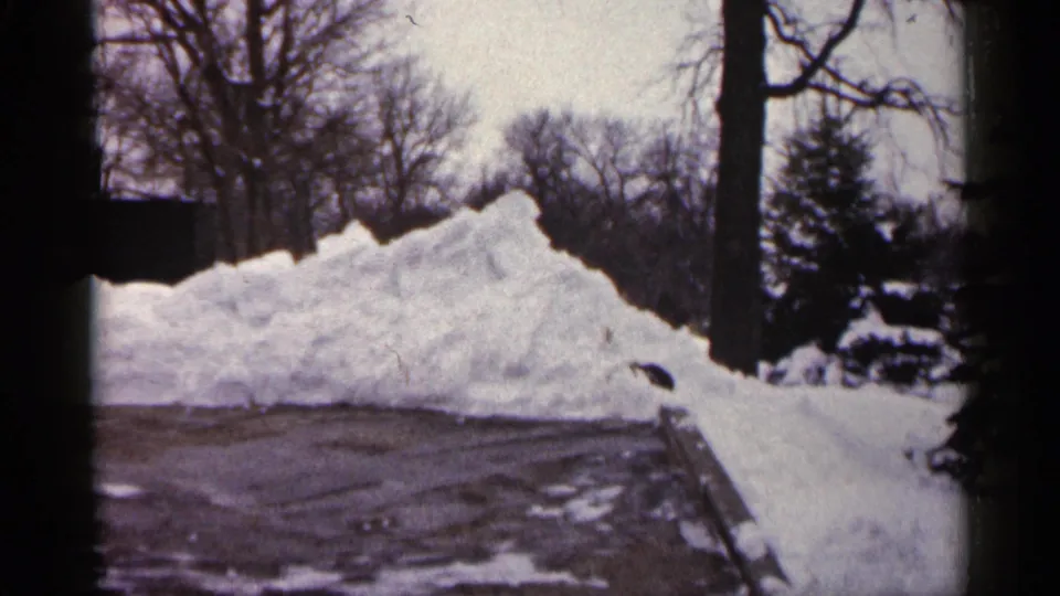 1961 Minnesota home-movie view of a high snowbank along wet pavement, framed by leafless trees and soft 8mm film edges.