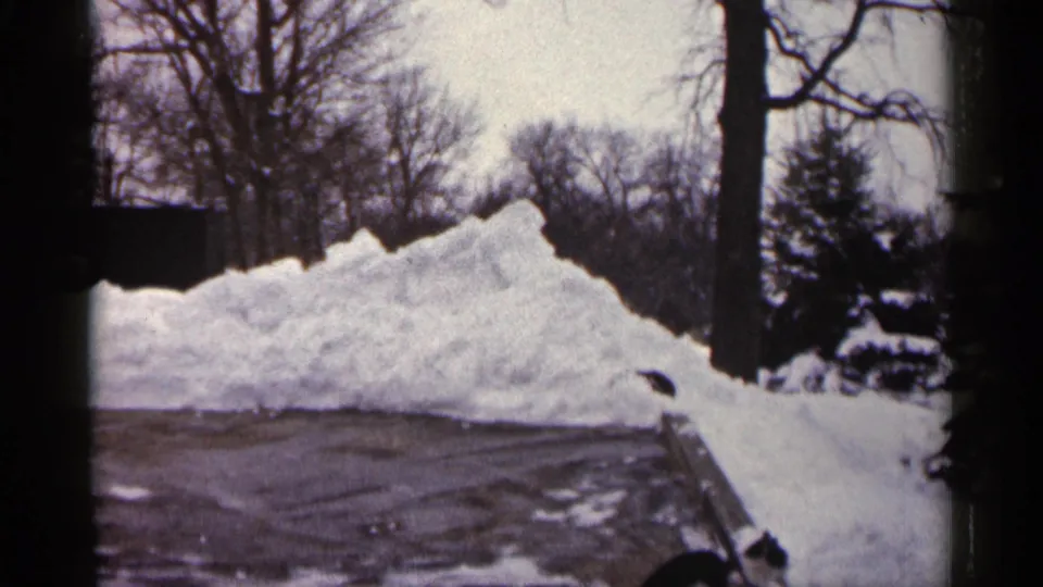 1961 Minnesota home-movie frame showing a large snowbank along a dark driveway with bare trees beyond it.