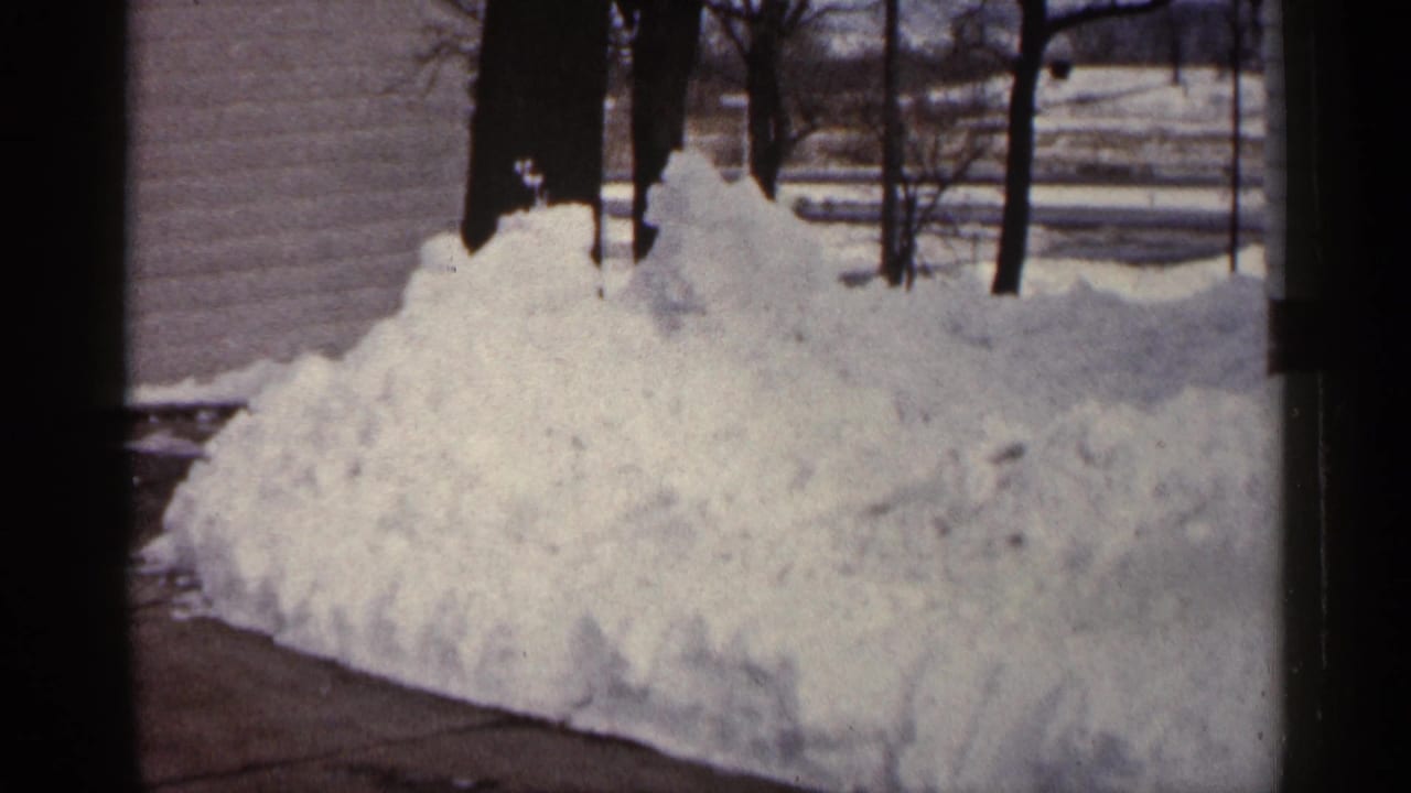1961 Minnesota frame with a piled snowbank beside driveway pavement, a house wall, and leafless trees.