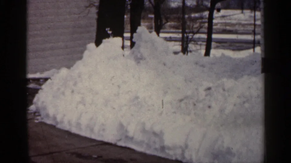 1961 Minnesota home-movie frame showing a mound of plowed snow beside a driveway with bare trees behind it.