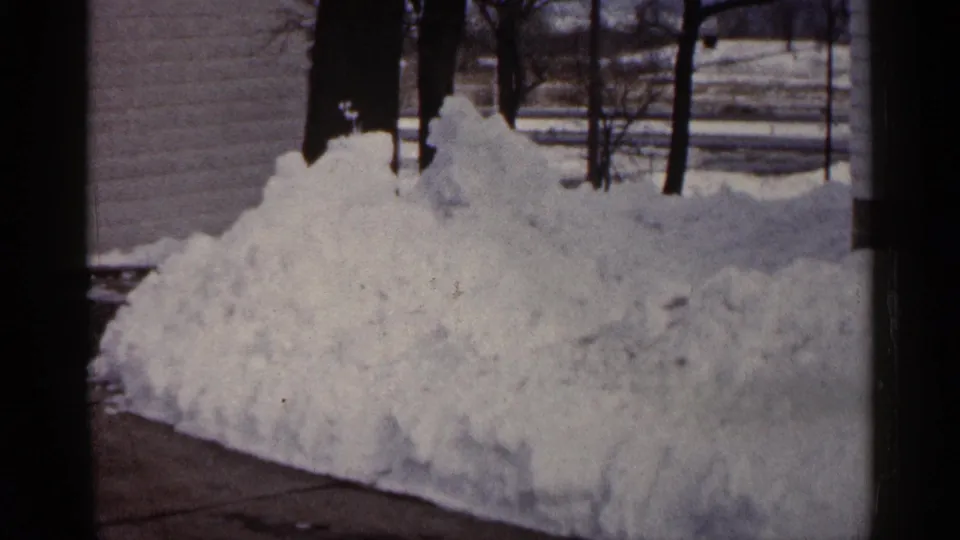 1961 Minnesota winter frame showing a residential snowbank, driveway edge, house wall, and leafless trees.