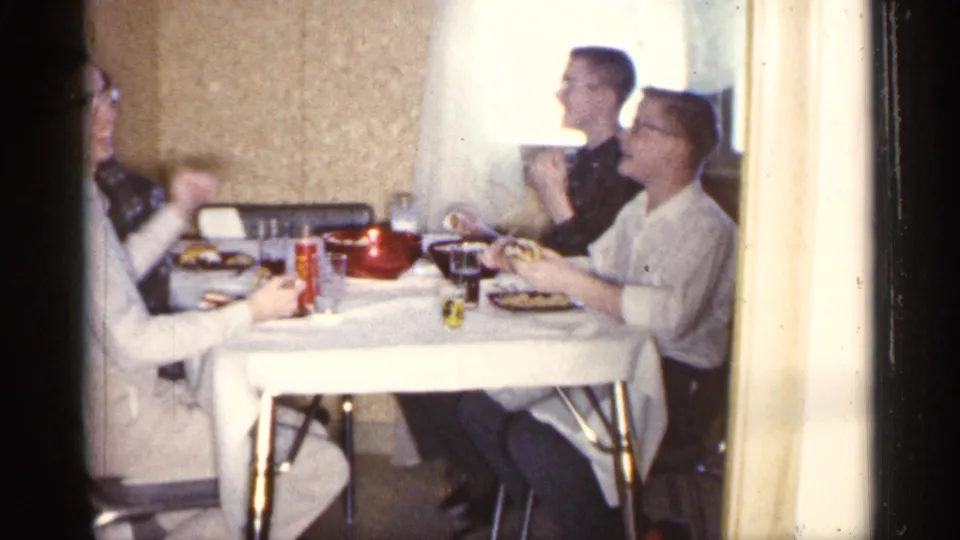 Three men sit at a dining table in a 1961 Minnesota home interior, with plates, glasses, a red pot, and pale curtains visible.