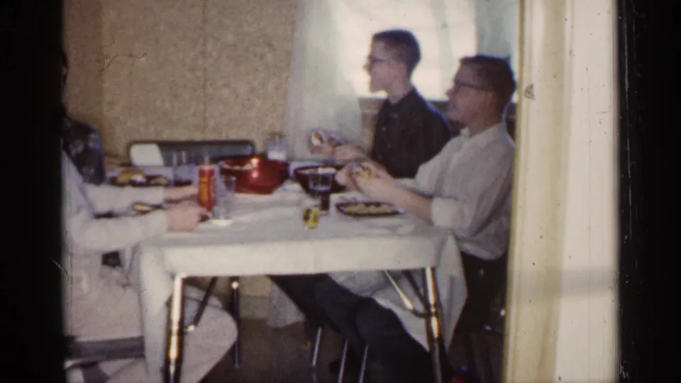 Two men are clearly visible at a dining table in a 1961 Minnesota home interior, with a third person partly at frame edge and tableware in front of them.