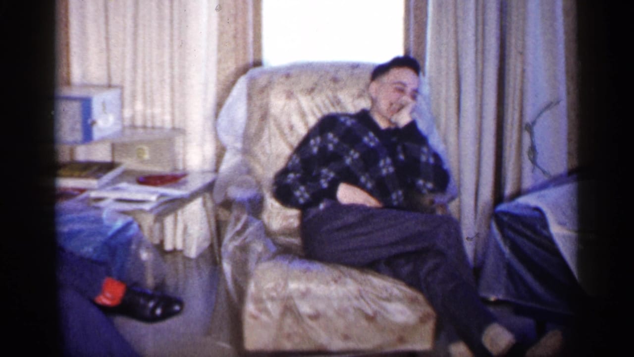 In a 1961 Minnesota home interior, a young man in a dark plaid sweater laughs while seated in a floral armchair.