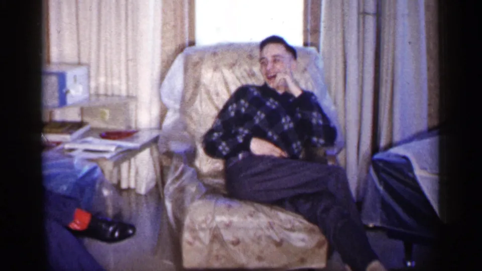 A young man reclines in a floral armchair in a 1961 Minnesota living room, smiling with his hand near his face.