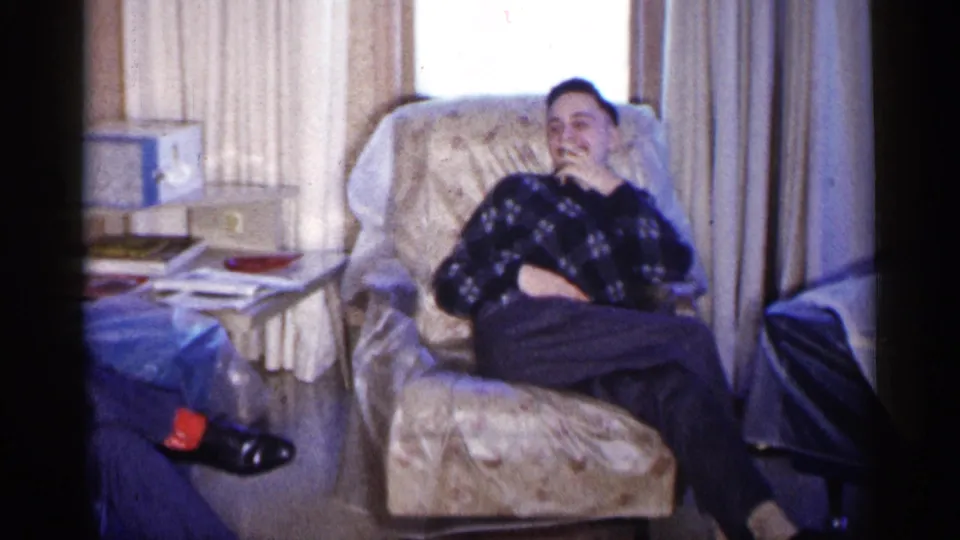 The 1961 Minnesota home-movie frame shows a young man seated in a floral armchair, partly covering his mouth.