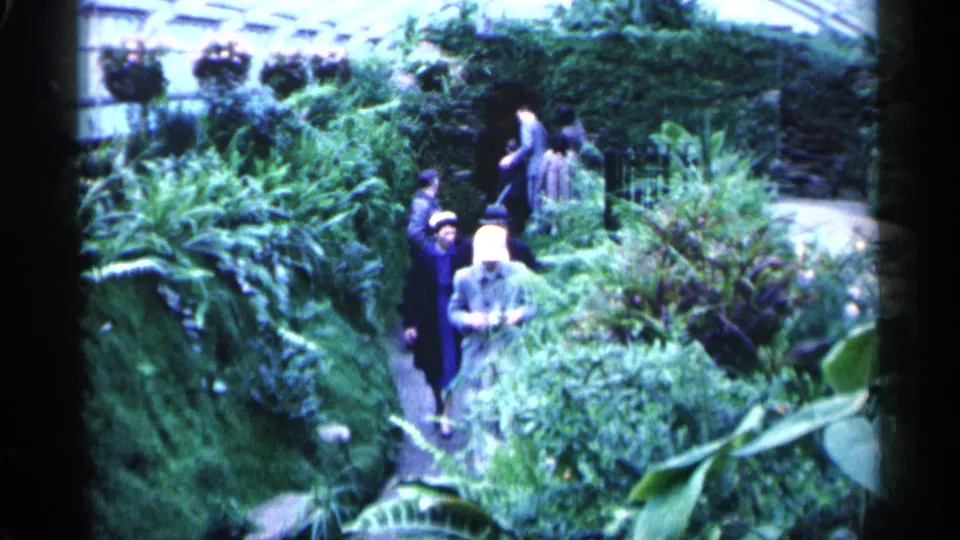 1961 Minnesota home-movie frame with visitors in coats and hats walking through a lush greenhouse garden path.