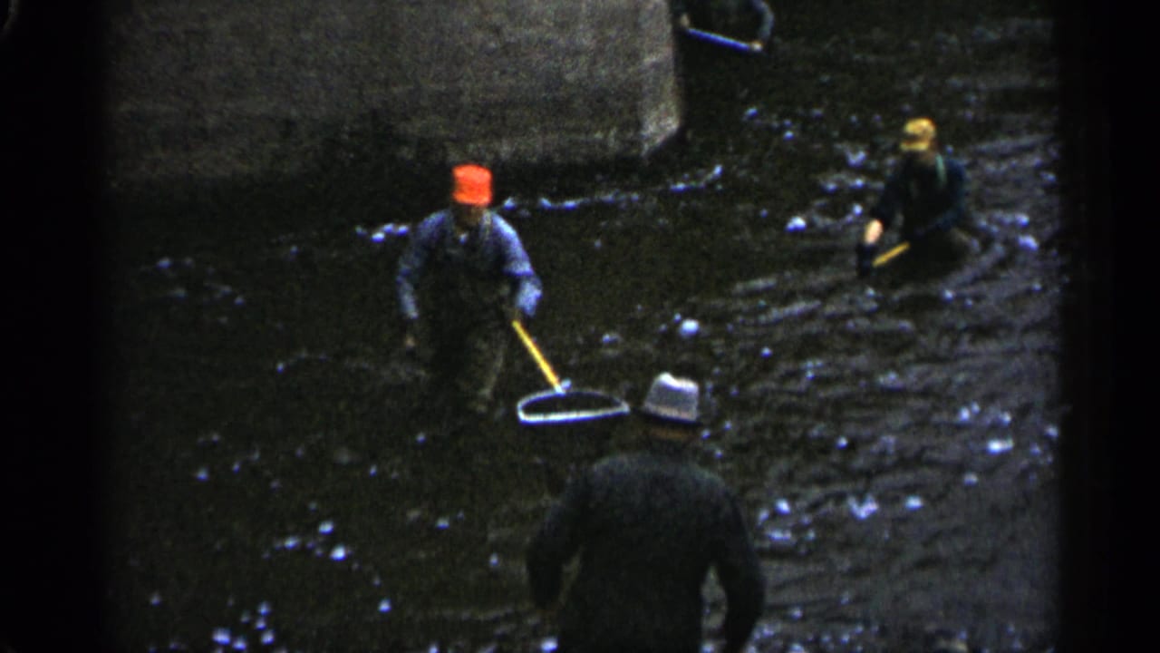 A man in a red cap holds a net while other men stand in the river behind him.