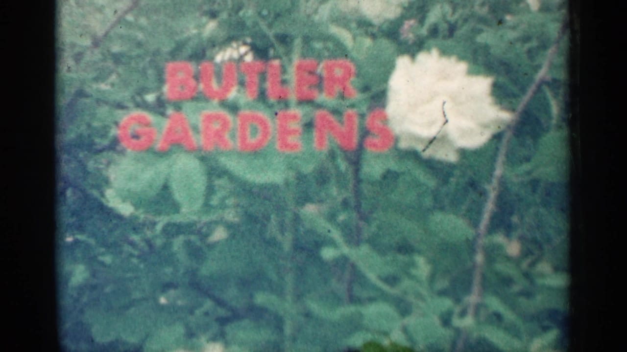 Red Butler Gardens lettering over green foliage with a white flower nearby.