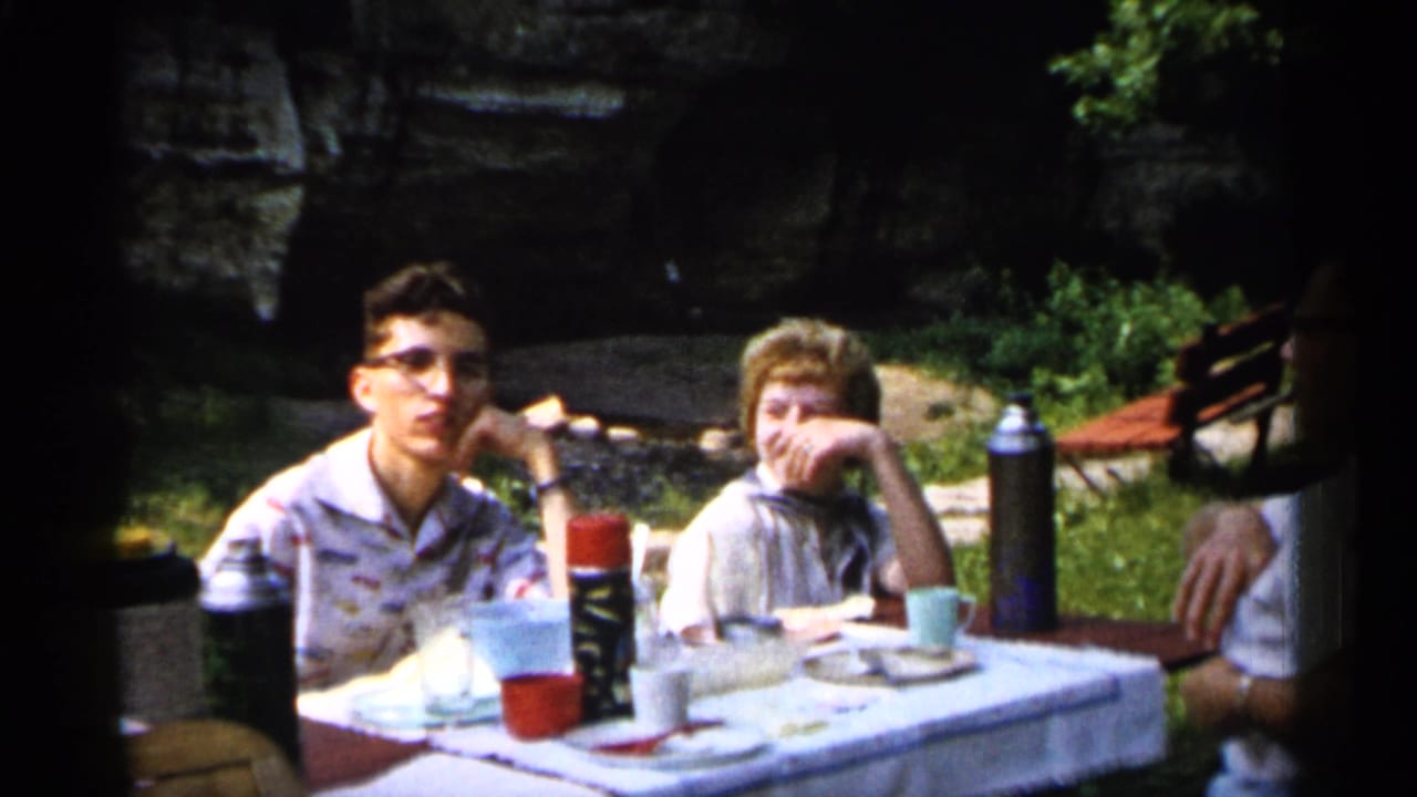 1961 Minnesota home-movie frame showing two family members at a cloth-covered picnic table in a park with rocks and foliage behind them.