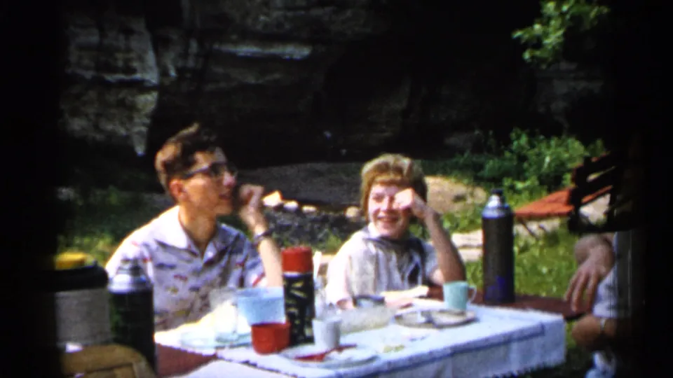 1961 Minnesota picnic scene with two smiling family members seated by tableware and thermoses, with a partial figure at the edge.