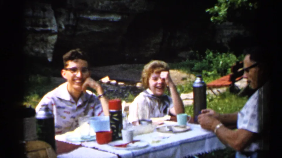 1961 Minnesota home-movie frame showing three adults around a picnic table, with the man at left smiling toward the camera.