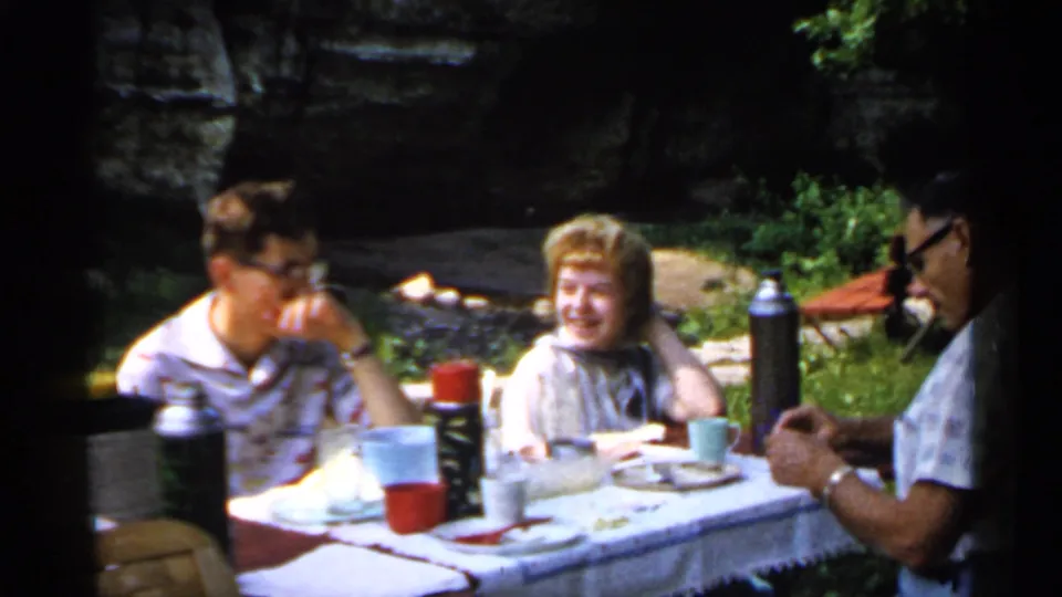 1961 Minnesota picnic frame showing adults seated around a cloth-covered table, with food items and thermoses in a park setting.