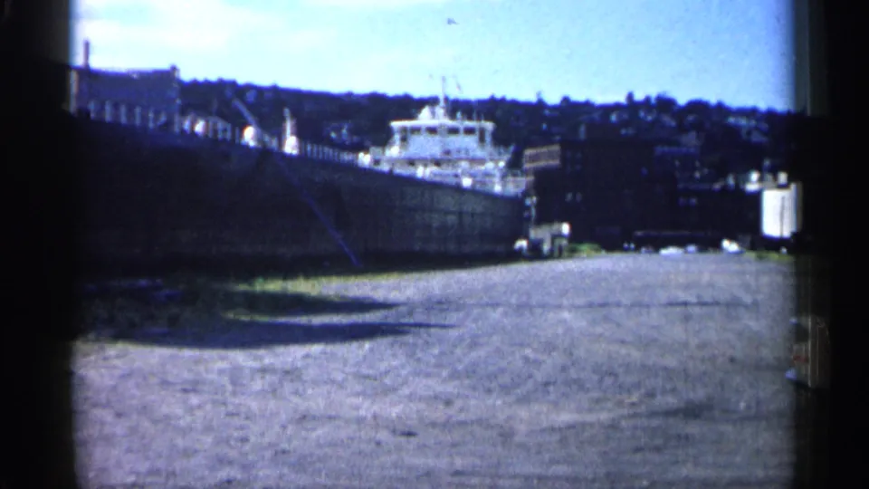 1961 Minnesota 8mm harbor frame with a large ship moored near a dock, water in the foreground, and shoreline structures behind.