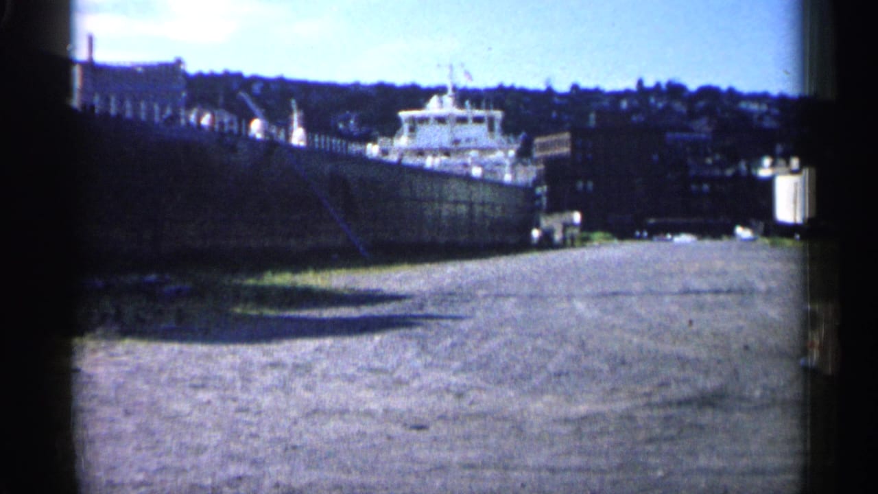 Wide view of a large docked ship beside a gravel shoreline and industrial waterfront.