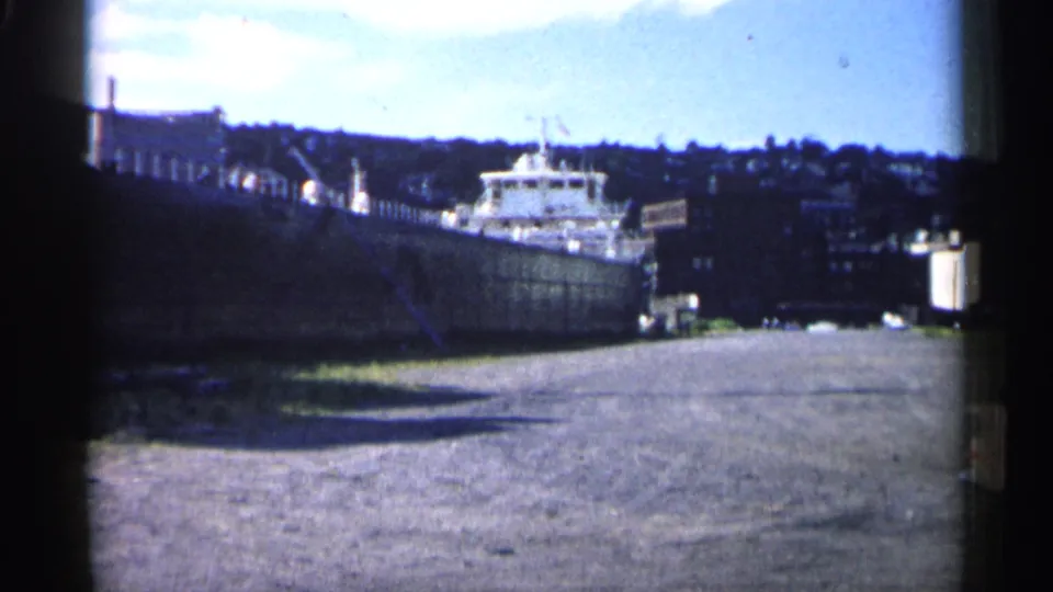 1961 Minnesota 8mm view of the Northern Venture docked at port, seen from shore with industrial buildings and a distant hillside.