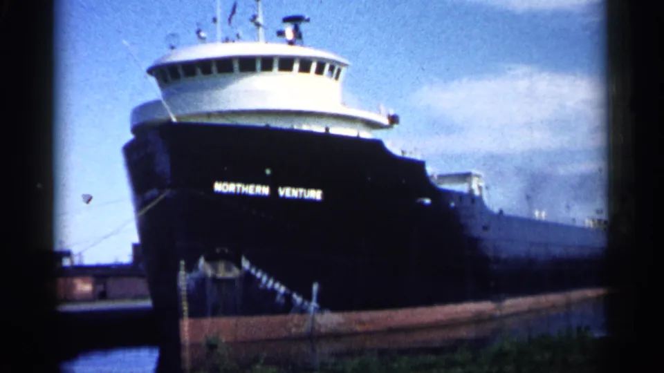 1961 Minnesota 8mm frame showing the Northern Venture bow, bridge windows, dock edge, and mooring line from a low shore angle.