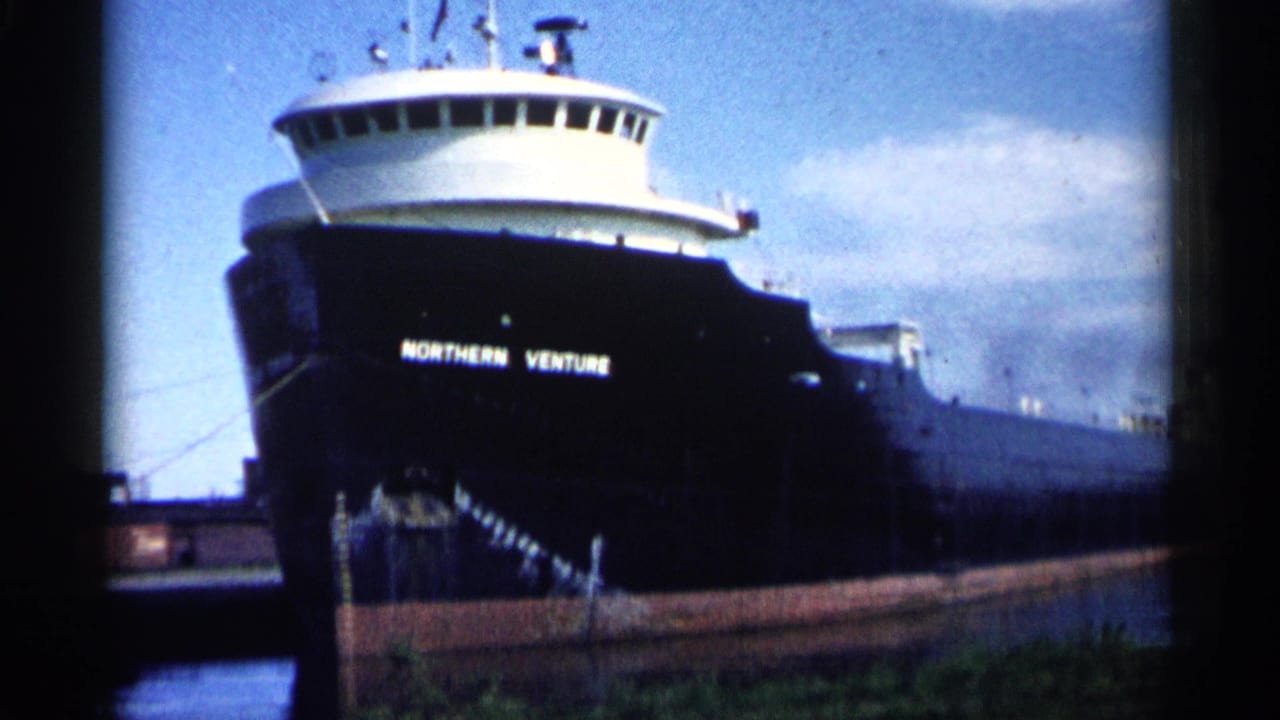 1961 Minnesota 8mm frame showing the Northern Venture freighter bow, white bridge, dark hull, and canal water under a blue sky.