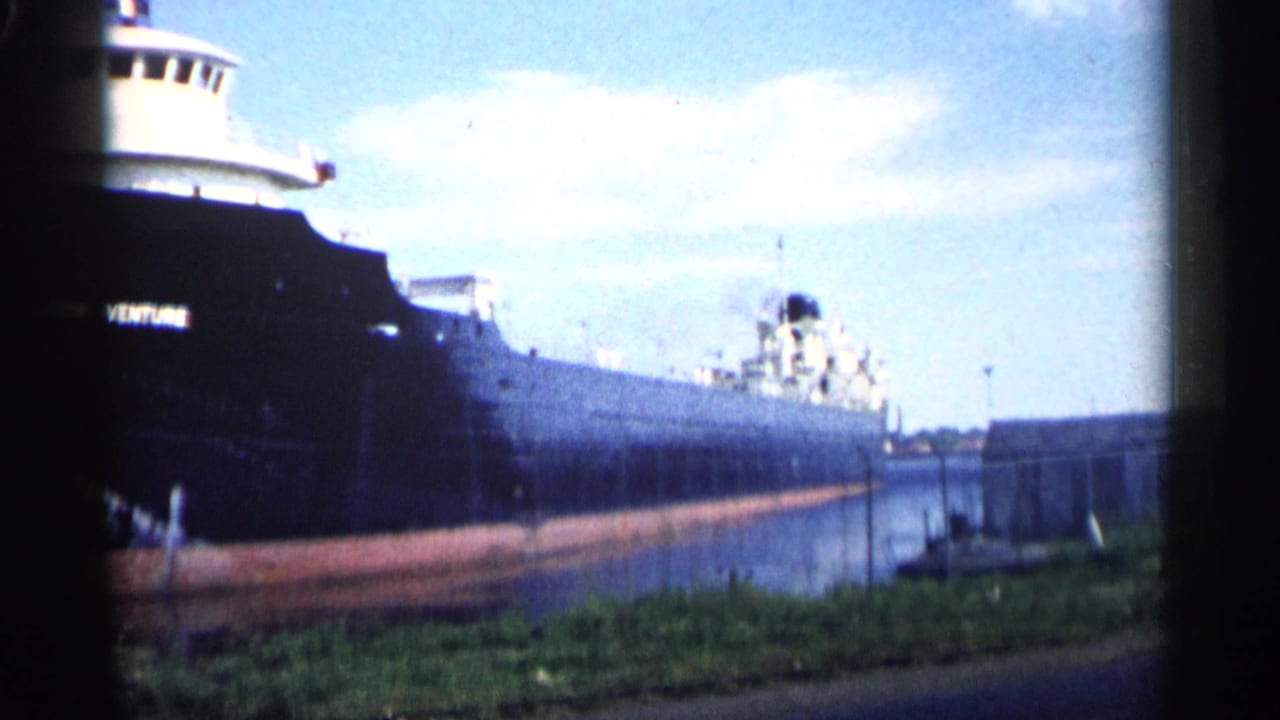 1961 Minnesota 8mm view of the Northern Venture stretching along a narrow waterway, with a grassy bank and industrial shoreline nearby.