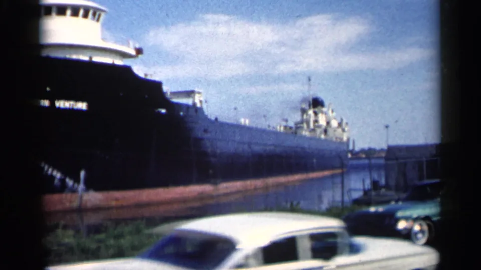 1961 Minnesota 8mm frame showing the Northern Venture in the canal while a white vintage car crosses the foreground near the shoreline.