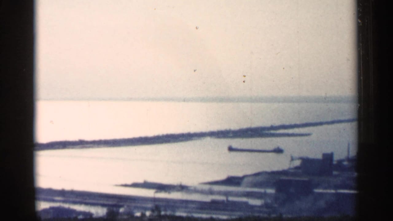Hazy harbor view with water, shoreline, distant barge, and industrial structures.