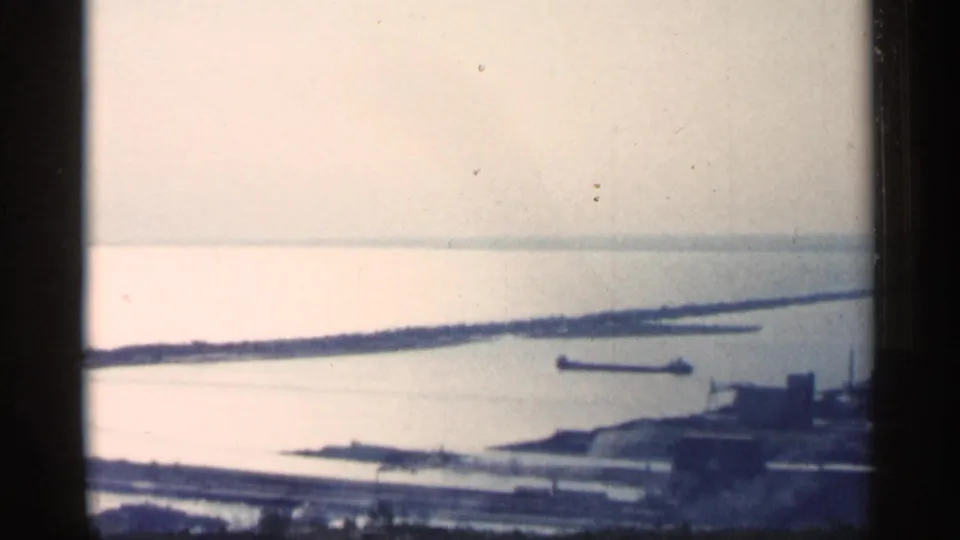 Wide hazy Minnesota harbor view from 1961 footage, with a distant barge on the water and low industrial shoreline structures.