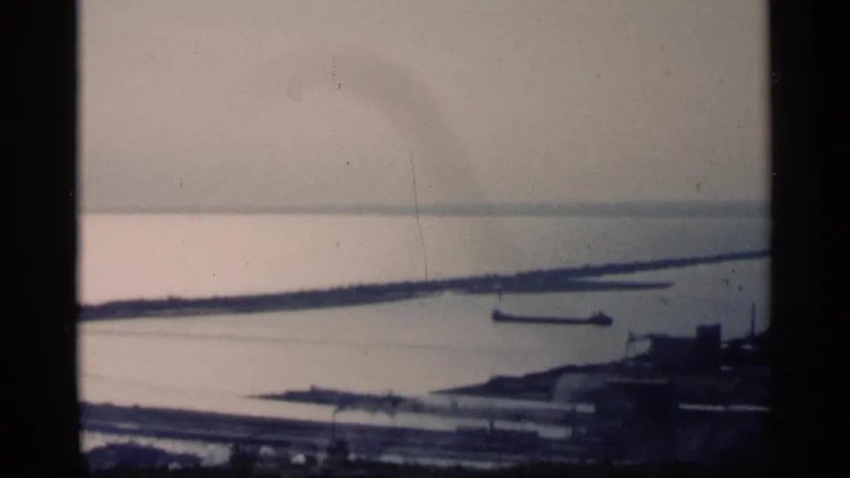 Final 1961 Minnesota harbor frame with a distant barge on calm water, shoreline bands, and dark industrial forms near the lower right.