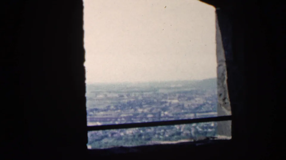 1961 Minnesota 8mm view through a stone window, showing a horizontal bar, distant city blocks, and possible water or shoreline below.