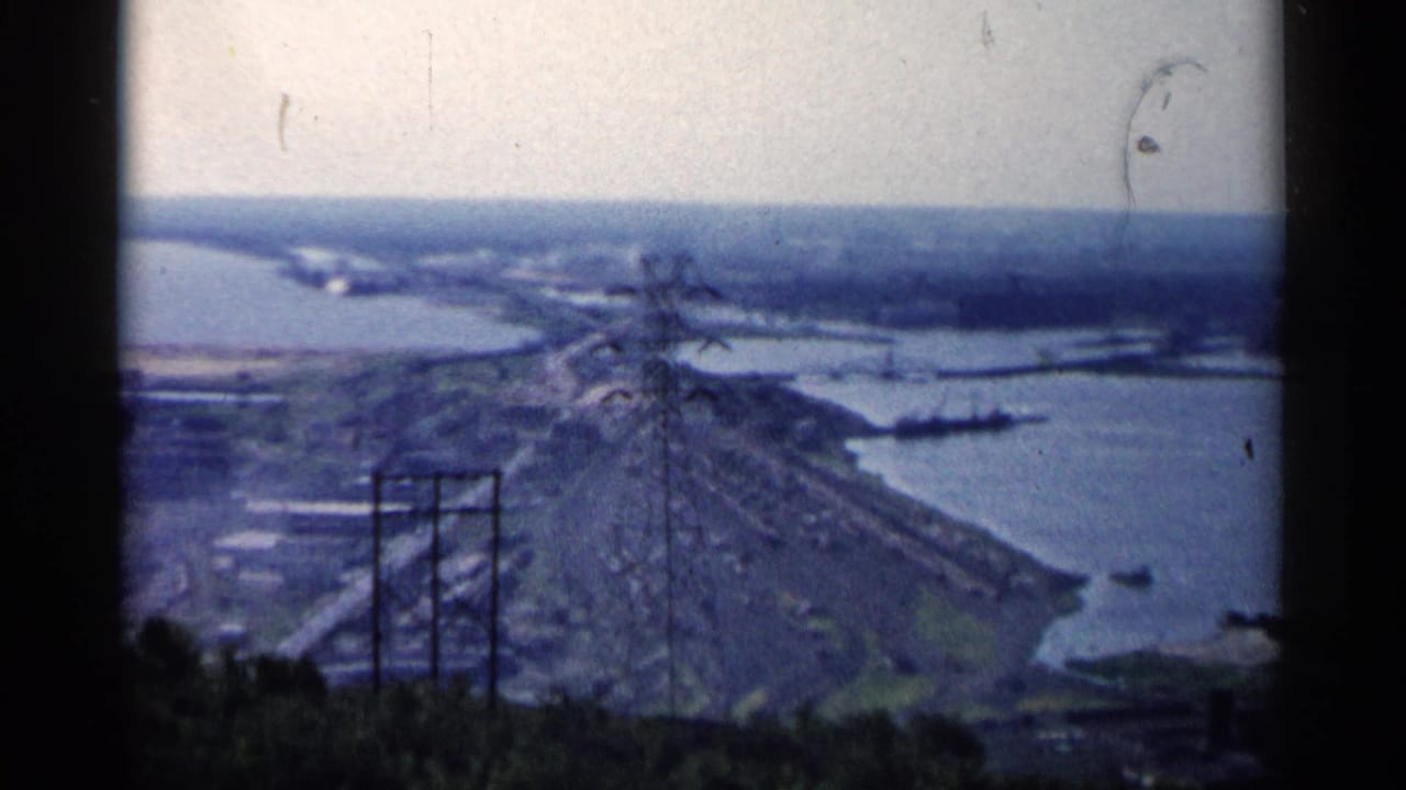 Elevated riverfront view with utility towers, shoreline, and distant city landscape.