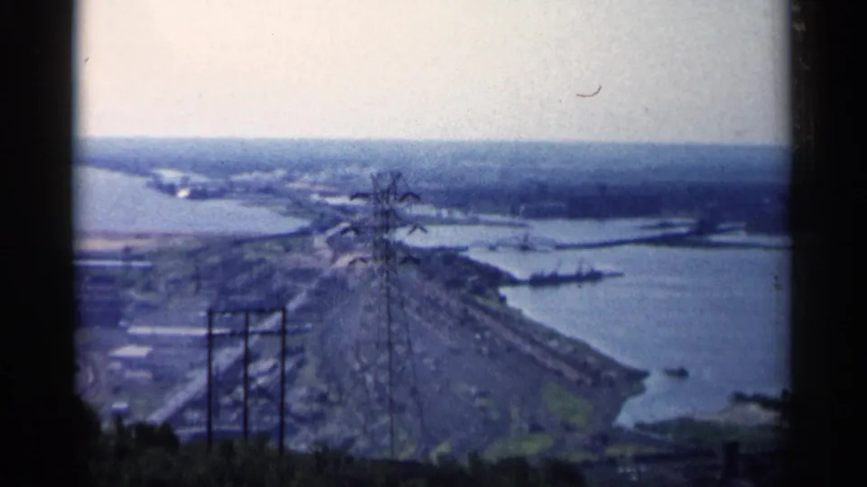 1961 Minnesota 8mm view over a riverfront corridor with utility structures, shoreline development, and a distant waterway under a pale sky.