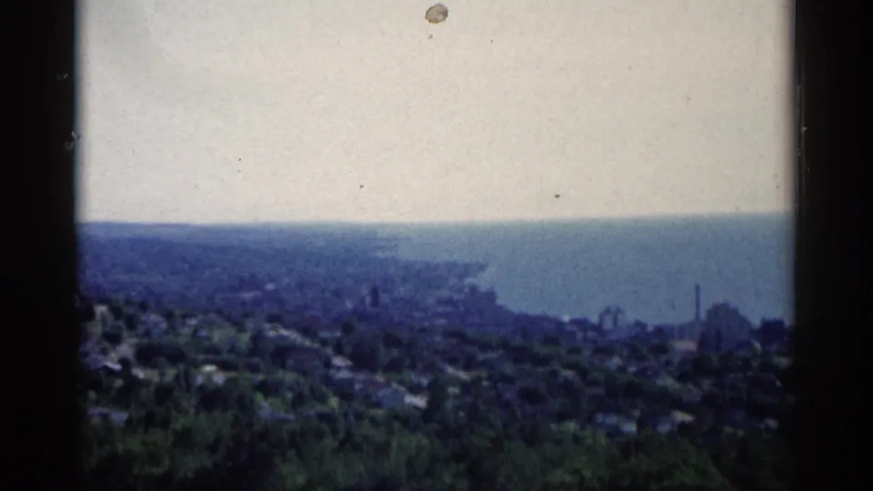 1961 Minnesota home-movie frame looking over a green hillside toward a distant city, waterfront horizon, and industrial silhouettes.