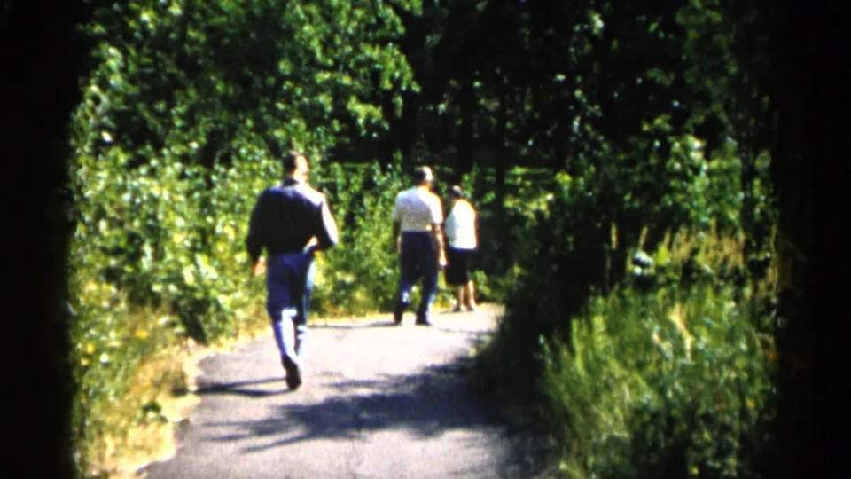 1961 Minnesota home-movie frame of three people walking away along a shaded green park path in summer light.