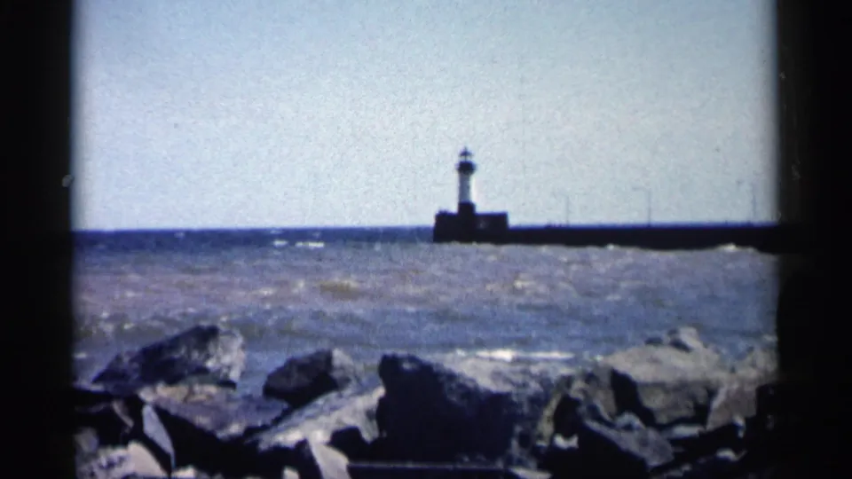 1961 Minnesota waterfront frame showing rocks in the foreground, rough water, and a lighthouse-like tower on a long pier.