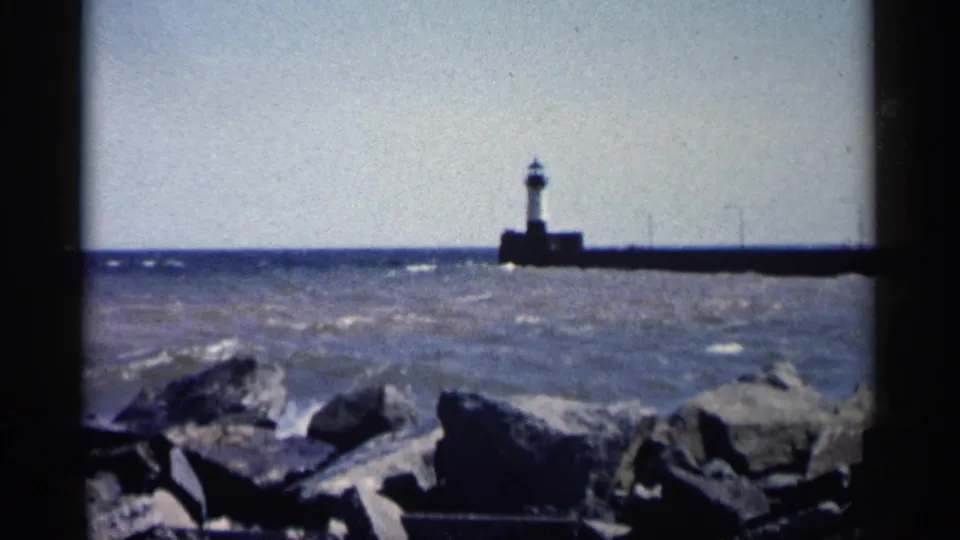 Vintage 8mm shoreline view with dark rocks, wind-ruffled water, and a small lighthouse tower at the end of a pier.