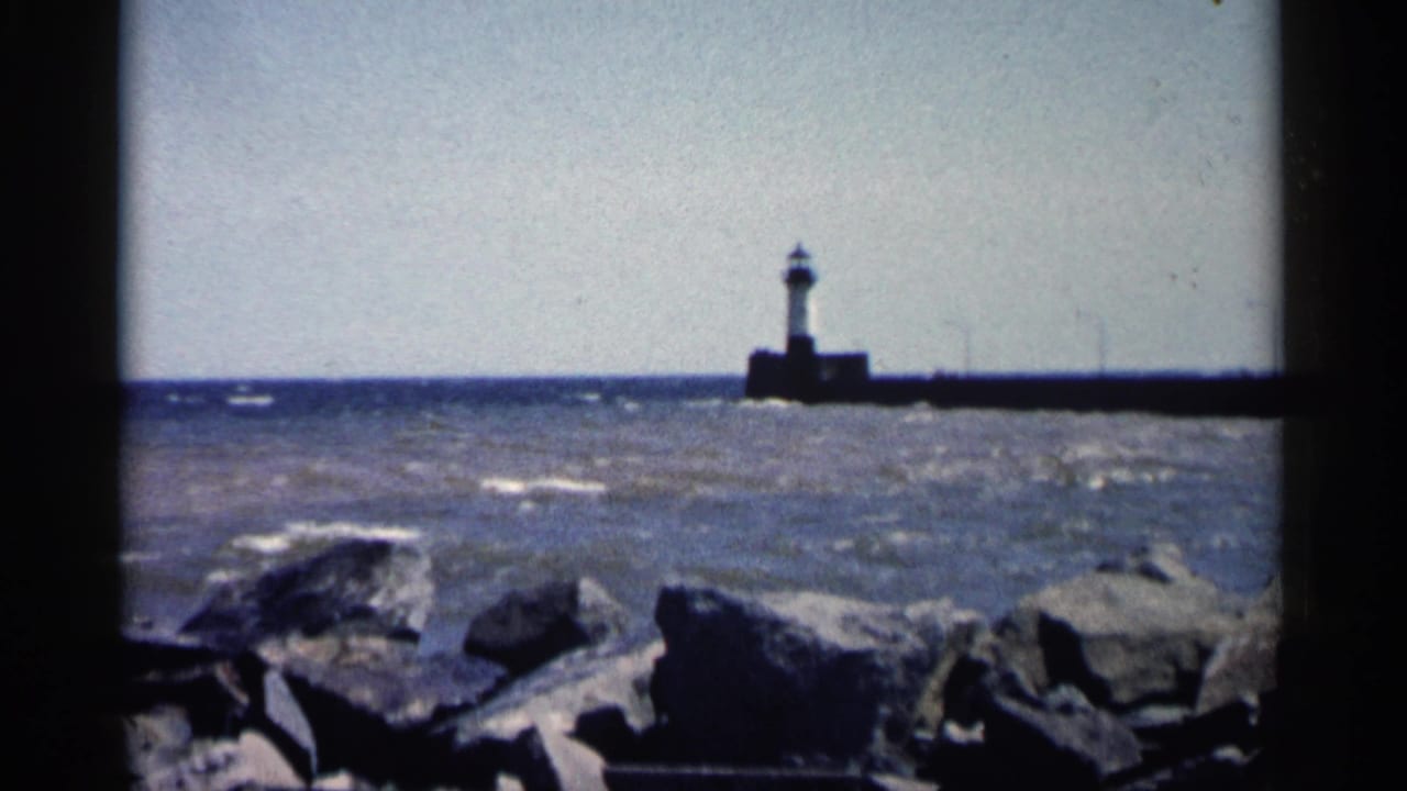 Distant lighthouse on a pier beyond choppy water and shoreline rocks.